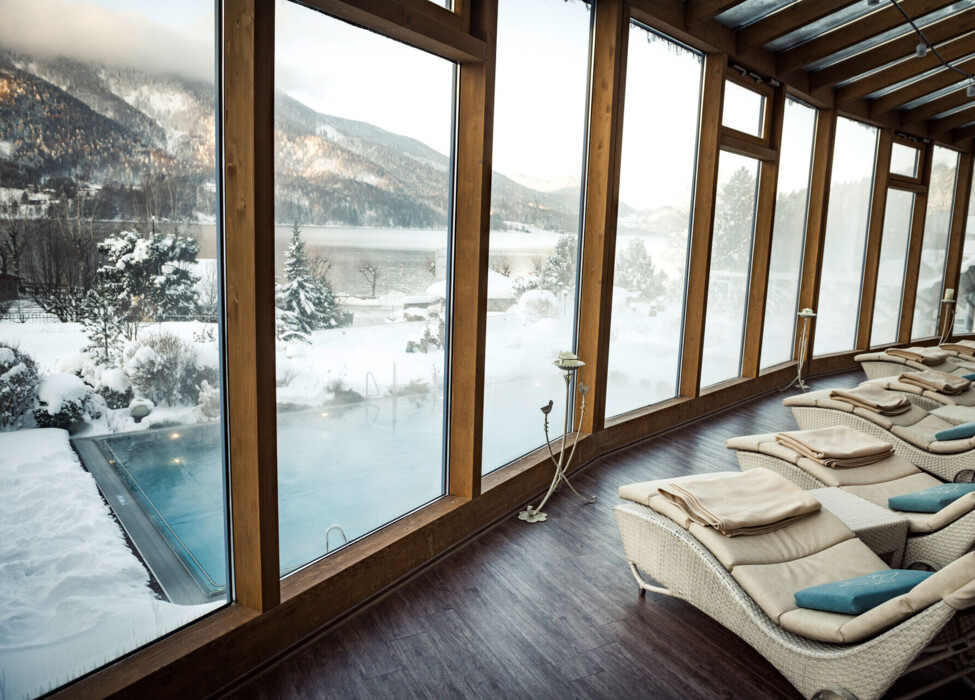 Lounge chairs by large windows overlooking a snow-covered scenic landscape with a pool and mountains in the background.