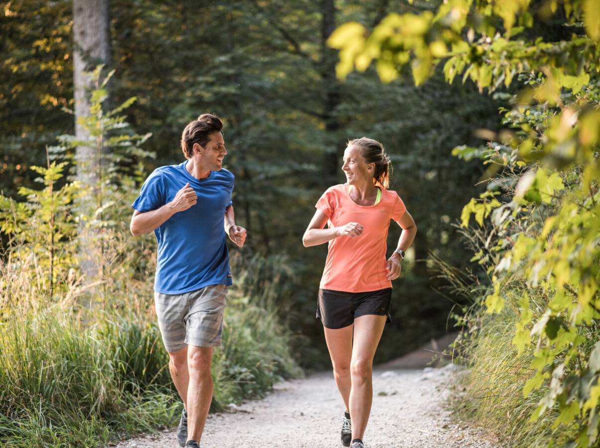 A man and woman jogging on a scenic forest trail, surrounded by lush greenery.