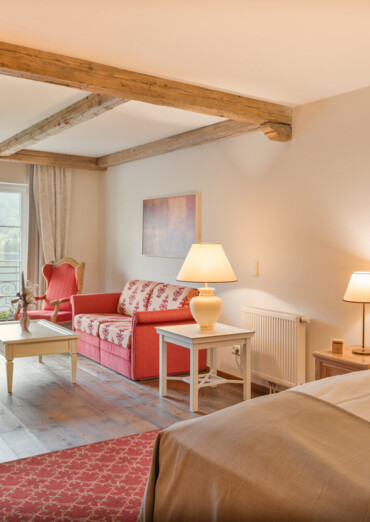 Cozy hotel room with wooden beams, a double bed, red sofa, and balcony doors. Warm lighting creates an inviting atmosphere.