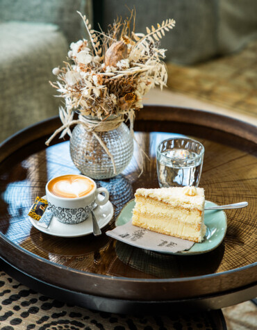 Kaffee und Kuchen auf einem Holztisch mit Trockenblumen in einem Glasgefäß im Hintergrund.