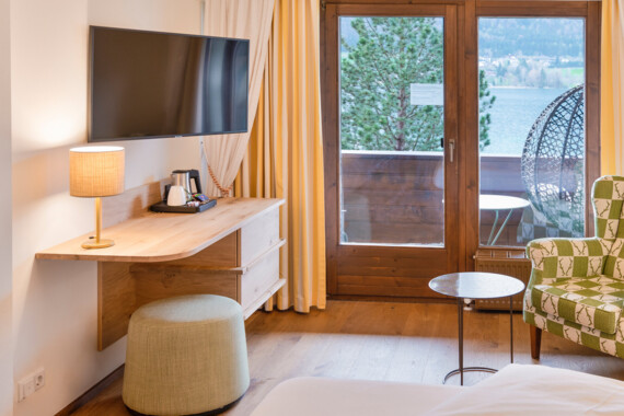Hotel room with a TV, wooden desk, and cozy chair, overlooking a balcony with a lake view at Waldhof Fuschlsee Resort.