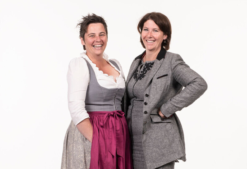 Two women smiling, one in traditional attire with a pink apron, and the other in a gray suit, standing together on a plain background.