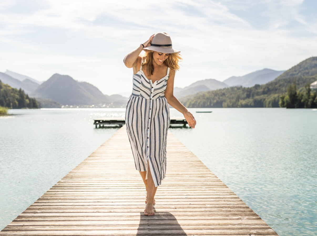 A woman in a striped dress and hat walks on a wooden pier over lake Fuschl, surrounded by lush mountains under a clear sky.