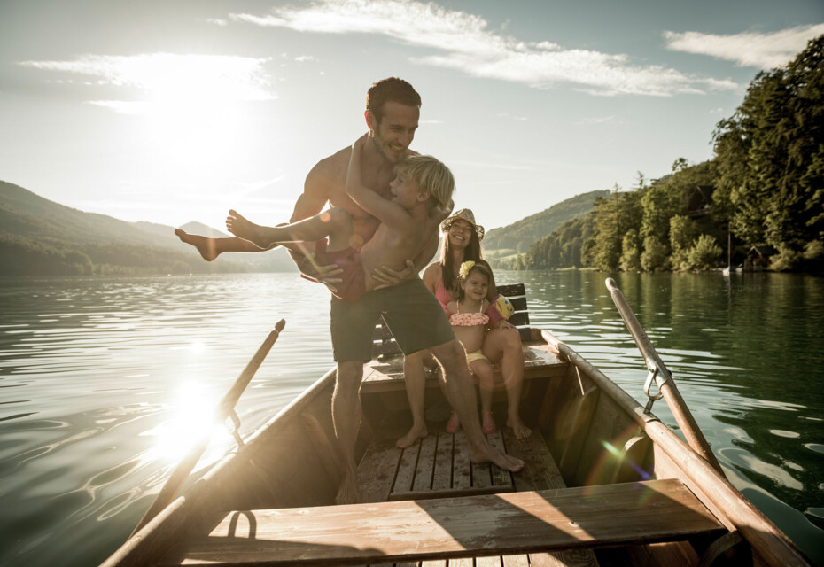 A family enjoying a boat ride on a tranquil lake surrounded by lush, scenic mountains and forests under the warm sunlight.