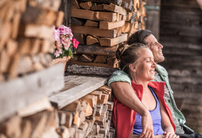 Ein Paar sitzt lächelnd an einer Hauswand, im Hintergrund sind aufgestapelte Holzscheite und ein Blumentopf mit rosa Blumen.