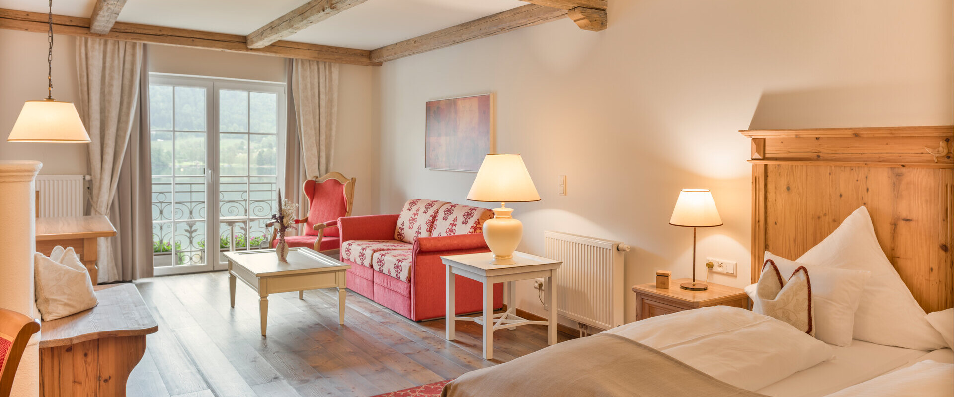 Cozy hotel room with wooden beams, a double bed, red sofa, and balcony doors. Warm lighting creates an inviting atmosphere.