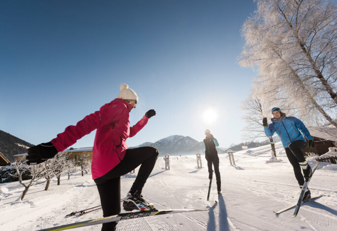 Three people cross-country skiing on a sunny winter day with mountains and trees in the background.