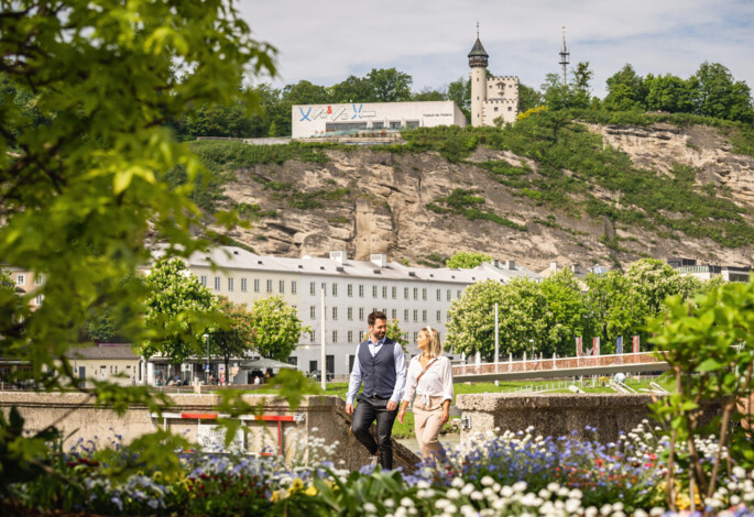 A couple walking along the Salzach river in Salzburg with a historic building on a hill in the background.