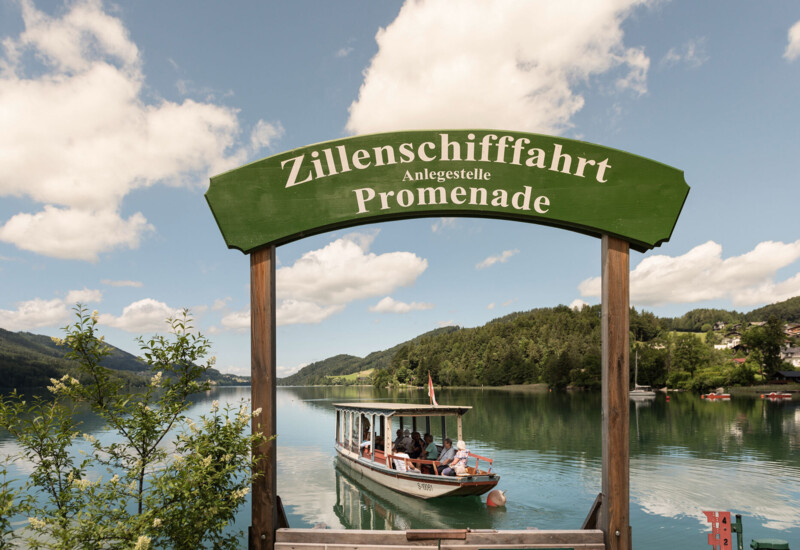 A boat departs from the Zillenschifffahrt promenade on a serene lake, surrounded by lush greenery and scenic hills under a partly cloudy sky.