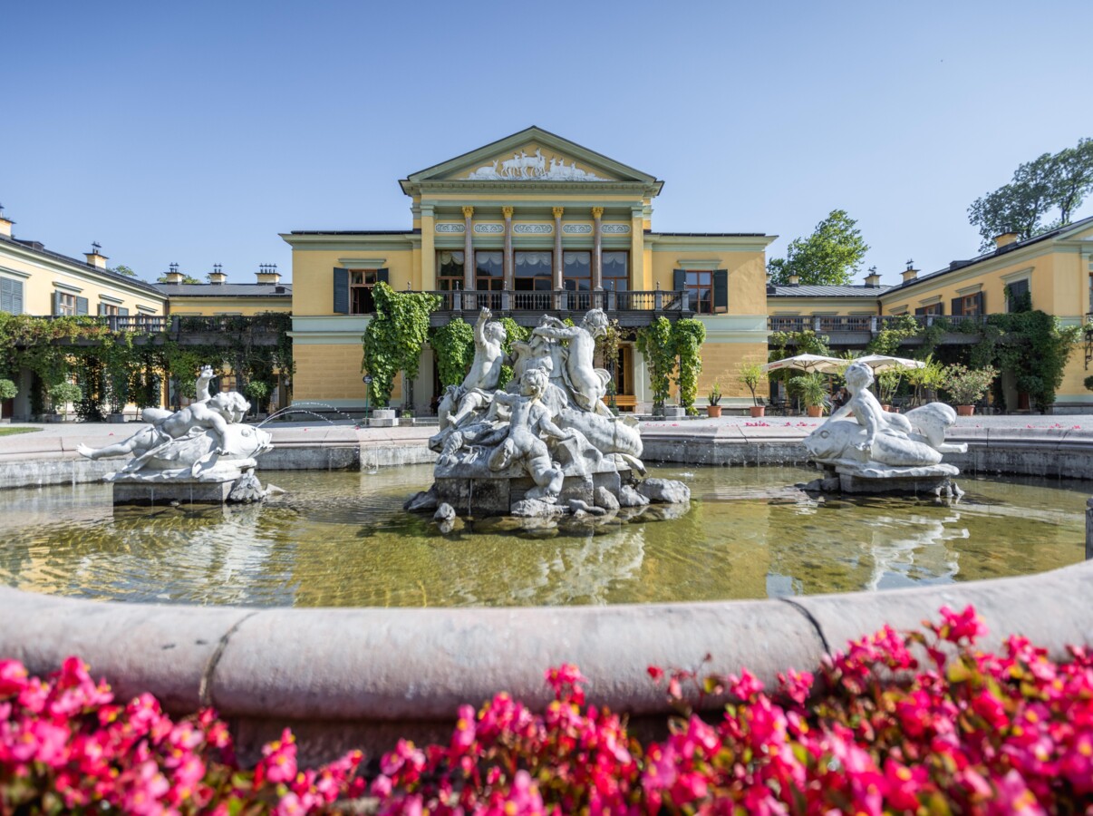 Fountain with statues in front of a grand building, surrounded by lush greenery and vibrant flowers, under a clear blue sky.
