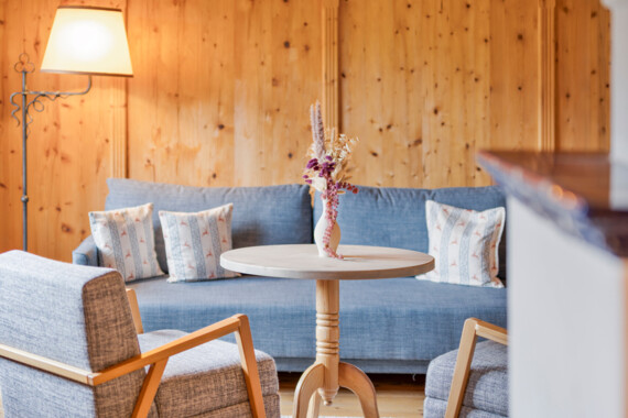 Cozy seating area with wooden walls, featuring a round table, two armchairs, and a blue sofa adorned with patterned cushions.