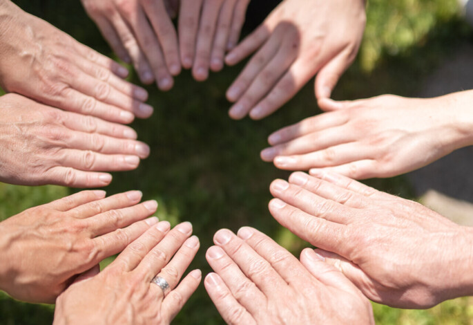 A circle of hands joined in unity on a grassy lawn.