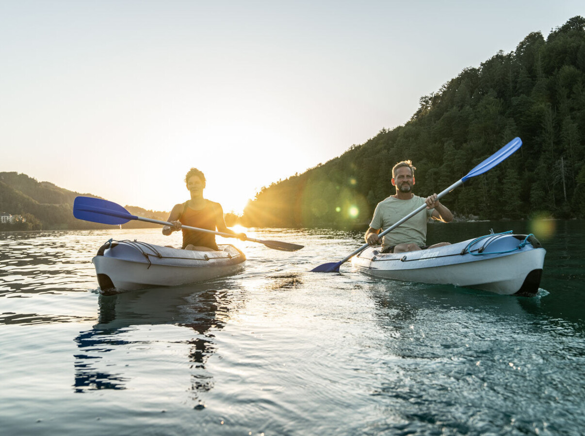 Two people kayaking on a calm lake at sunset, surrounded by lush, scenic forested hills.