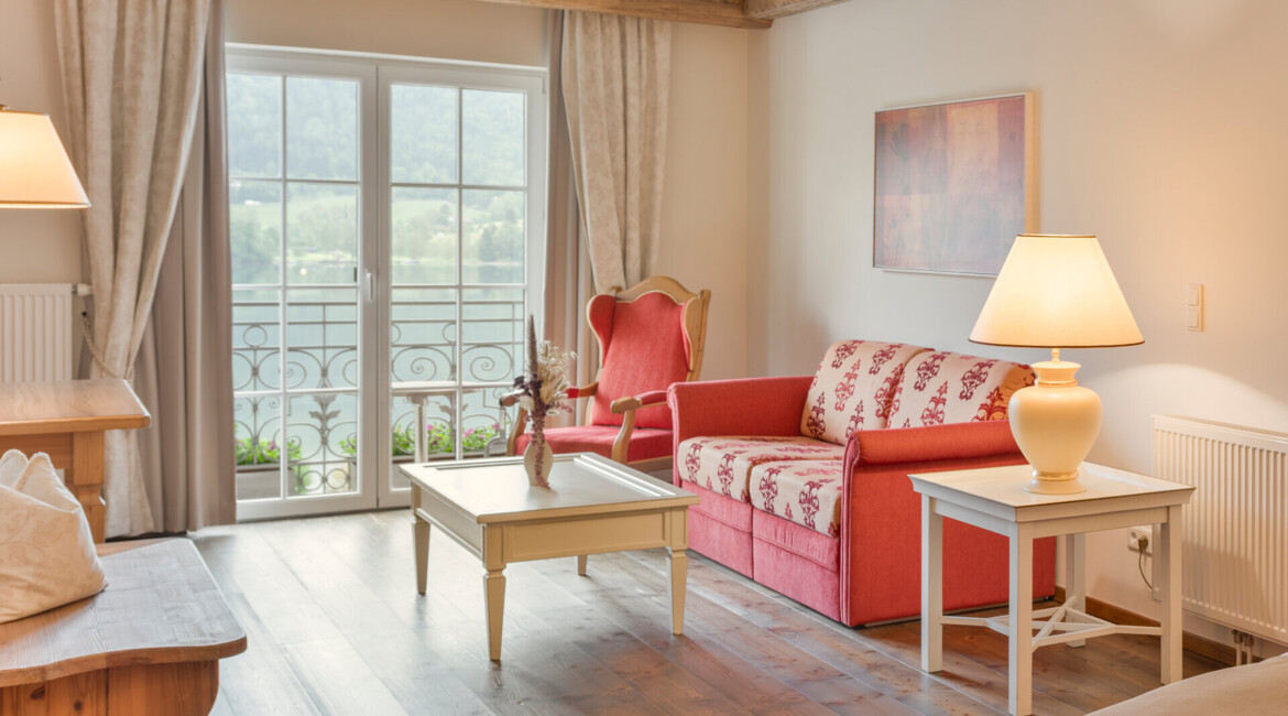 Cozy living room with elegant decor, featuring a red sofa, armchair, and wooden beams, overlooking a serene landscape through large windows.