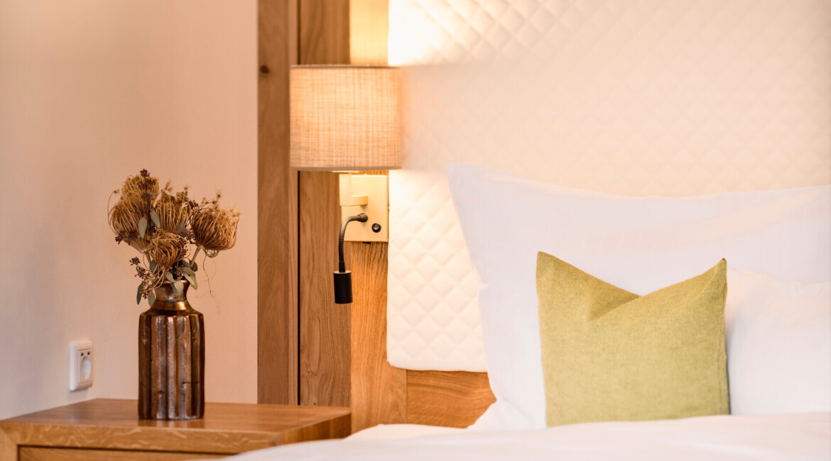 A cozy hotel room with a wooden headboard, white bedding, and a green pillow. A bedside table holds a vase with dried flowers, and a wall lamp provides soft lighting.