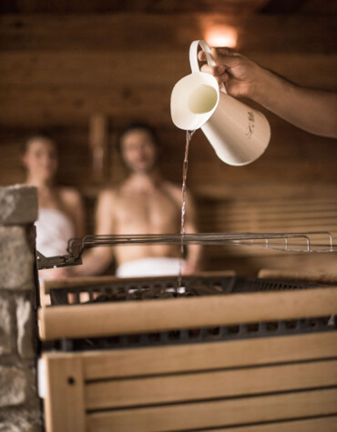 A person pours water over sauna stones, with two people in the background enjoying the warmth.