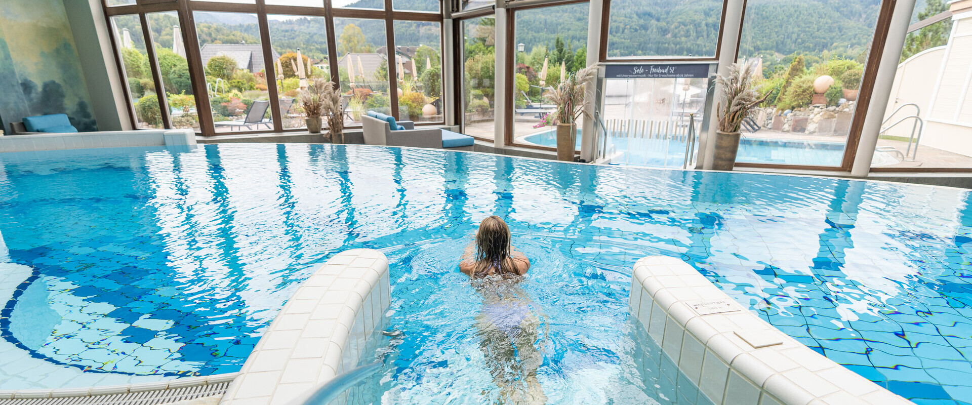 A person swims in an indoor pool with large windows at Waldhof Fuschlsee Resort, offering views of the surrounding landscape.