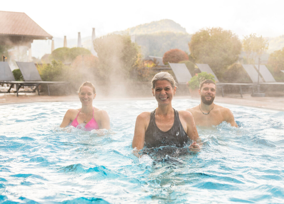 Three people enjoying a steaming outdoor pool, surrounded by lush greenery and scenic views.