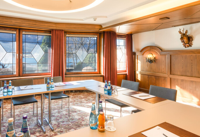 Meeting room with large windows and natural light, featuring a U-shaped table setup with bottled water and notepads.