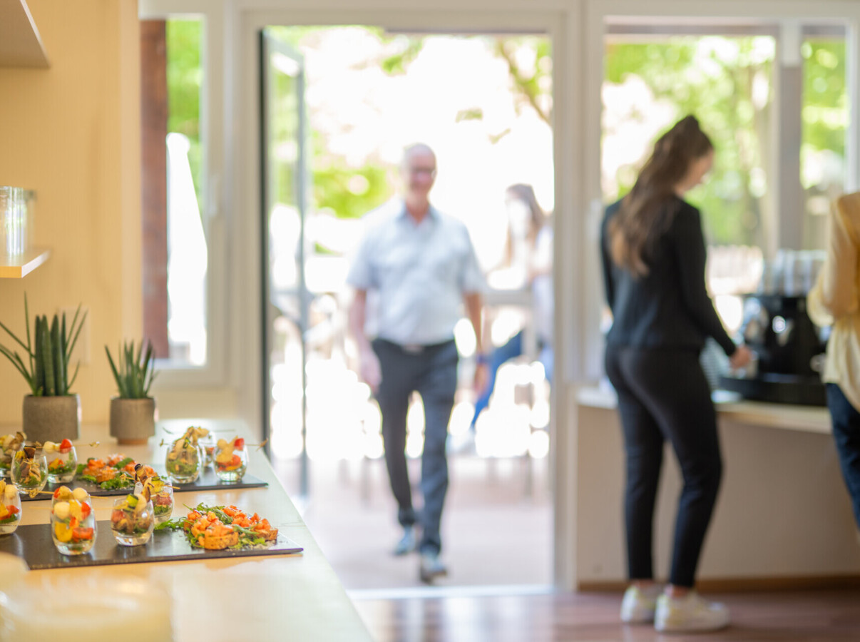 A table with appetizers and potted plants in a bright room, with people in the background near large windows.