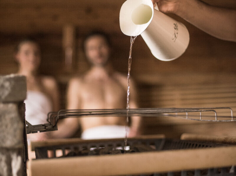 Water being poured onto sauna stones, creating steam, with two people in towels relaxing in the background.