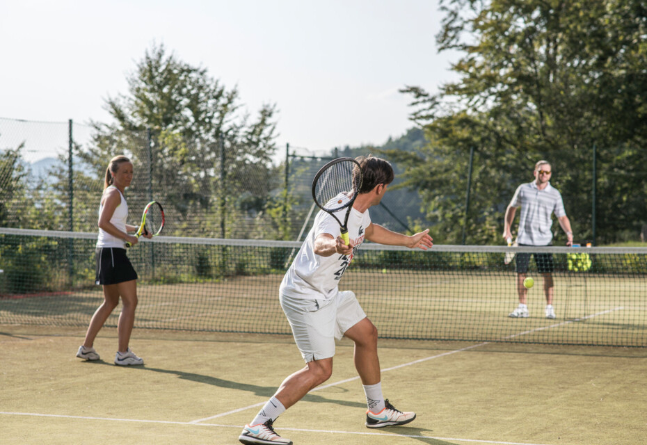 Three people playing doubles tennis on a scenic outdoor court surrounded by lush trees.