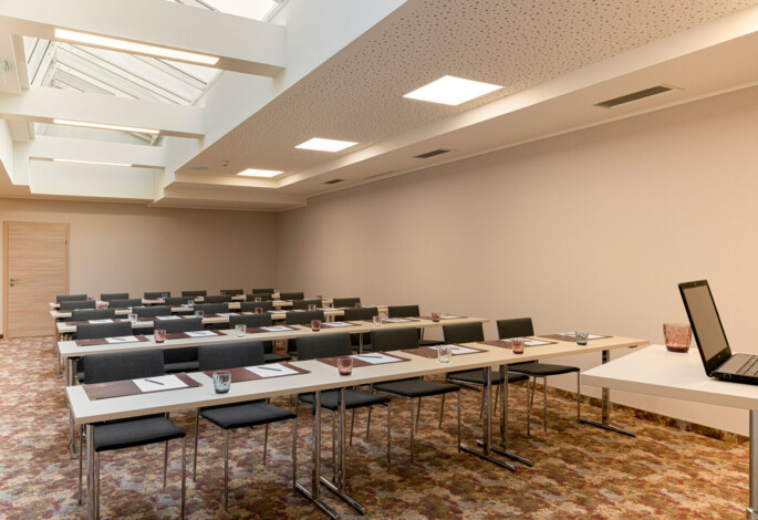 A well-lit conference room with rows of tables and chairs, featuring a skylight and a laptop on a desk.