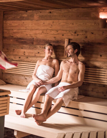 Personen entspannen sich in einer Sauna aus Holz; ein Mann wirbelt ein Handtuch für Luftzirkulation.