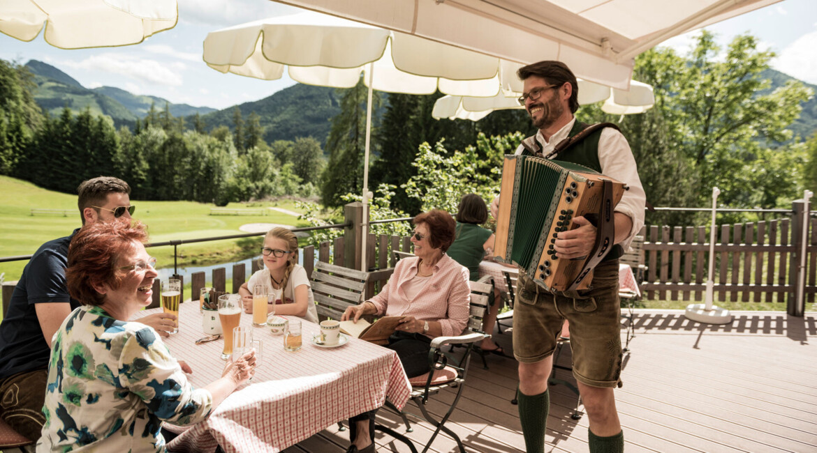 A man plays an accordion for a smiling family on a sunlit patio overlooking lush, scenic mountains.