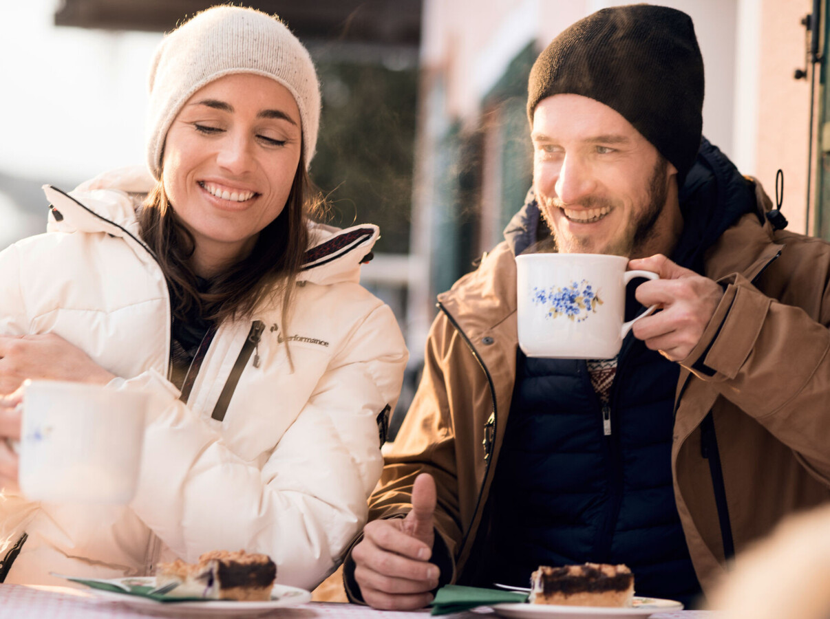 A couple enjoys hot drinks and cake outdoors in winter attire, smiling warmly.