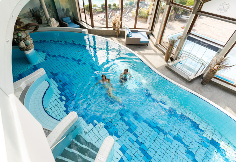 Indoor pool area with two people swimming, surrounded by large windows offering natural light and a view of the outdoors.