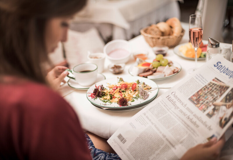 A person enjoys breakfast while reading a newspaper at Waldhof Fuschlsee Resort, featuring a variety of dishes, coffee, and a glass of juice.
