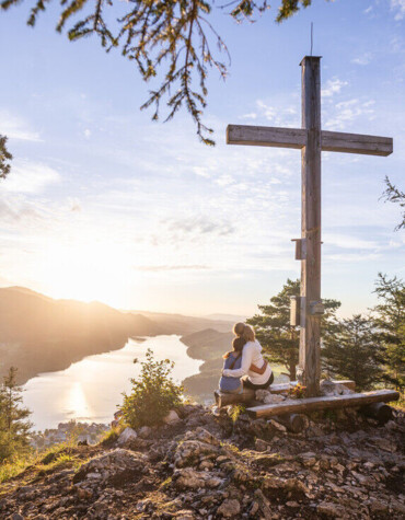 A woman and her daughter sit by a wooden cross on a mountain with a view of a lake and sunset.