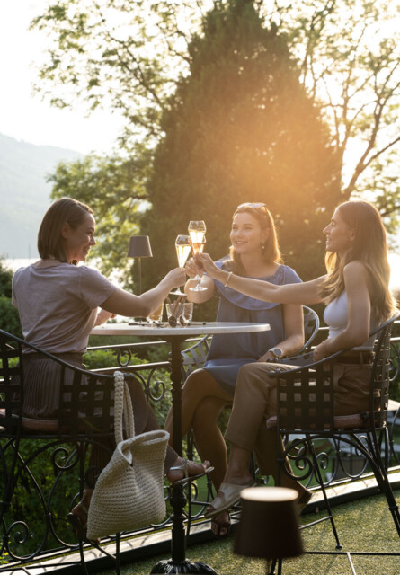 Three women toasting with drinks on a patio, surrounded by lush greenery and scenic views.