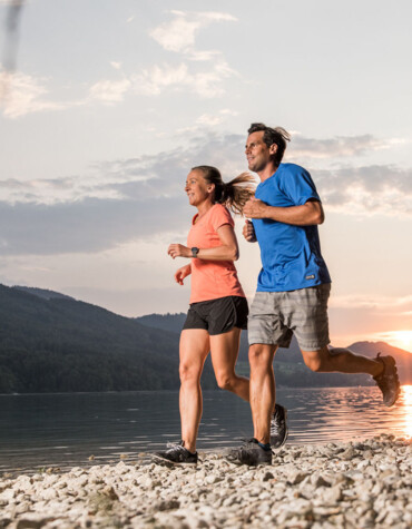 A couple jogging along a lakeside trail at sunset, with scenic mountains in the background.