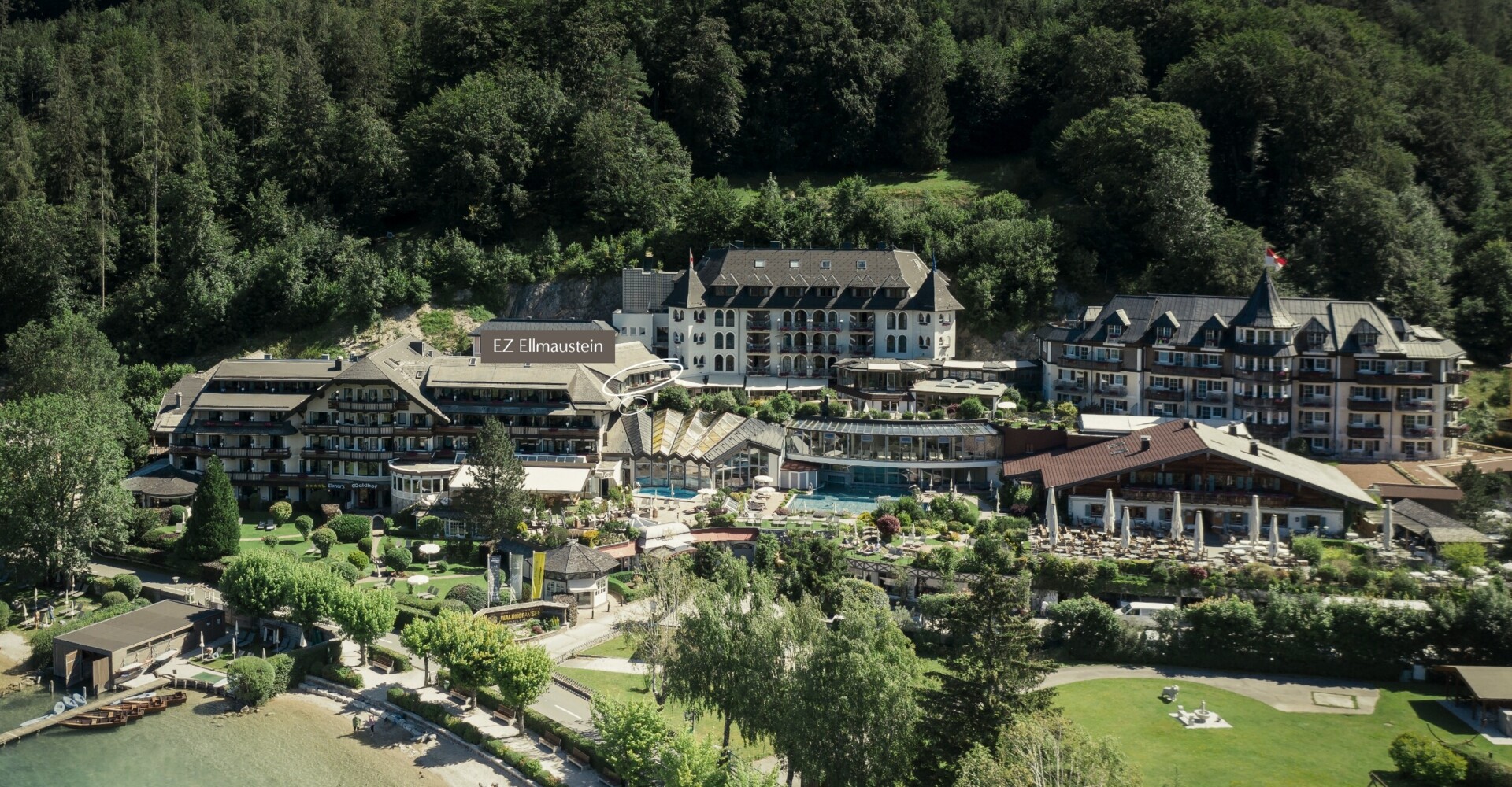 Luftaufnahme vom Waldhof Fuschlsee, umgeben von üppigen Bäumen und Grünflächen, mit direktem Zugang zum Strand.