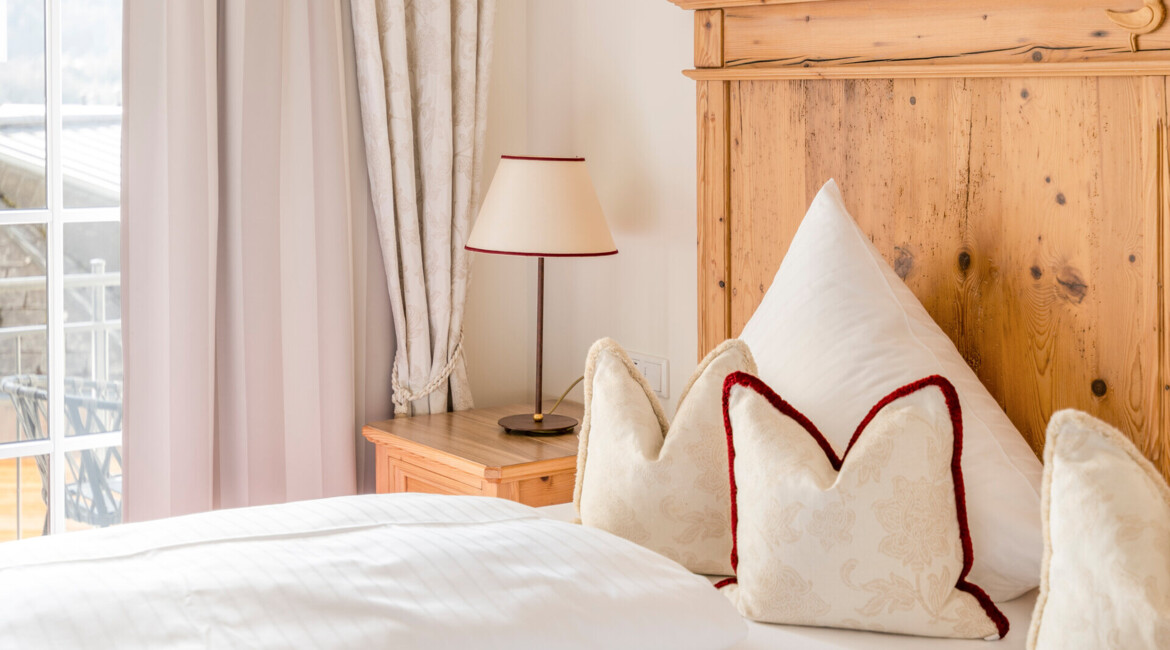 Bright, cozy hotel room with a wooden headboard, plump pillows, and a bedside lamp. Soft curtains frame a window with a scenic view.