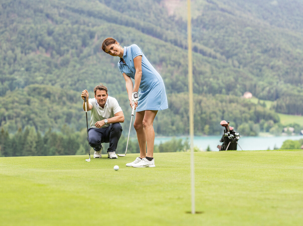 A woman puts on a golf course with a scenic, lush landscape in the background. A man crouches nearby, observing.