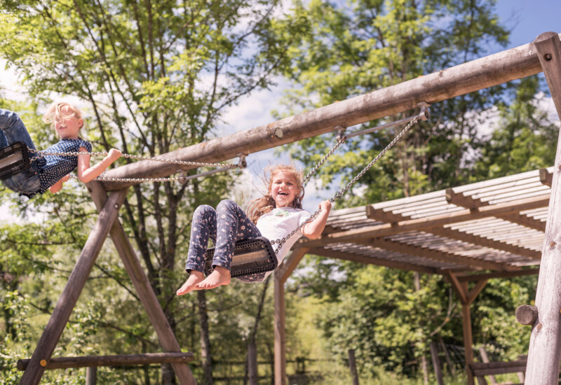 Kinder auf einer Schaukel am Outdoor-Spielplatz des Familienhotel Ebner's Waldhof am See.