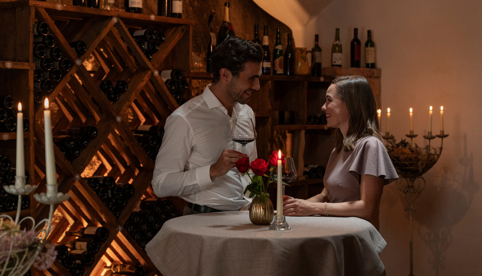 A couple enjoying wine in a cozy, candlelit wine cellar, surrounded by bottles on wooden racks.