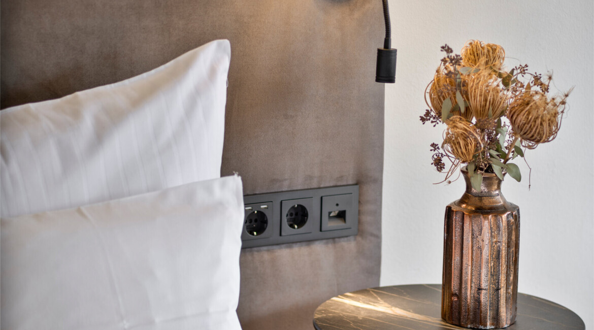 Bedside lamp with a soft light above a pillow, next to a vase of dried flowers on a table, creating a cozy atmosphere.