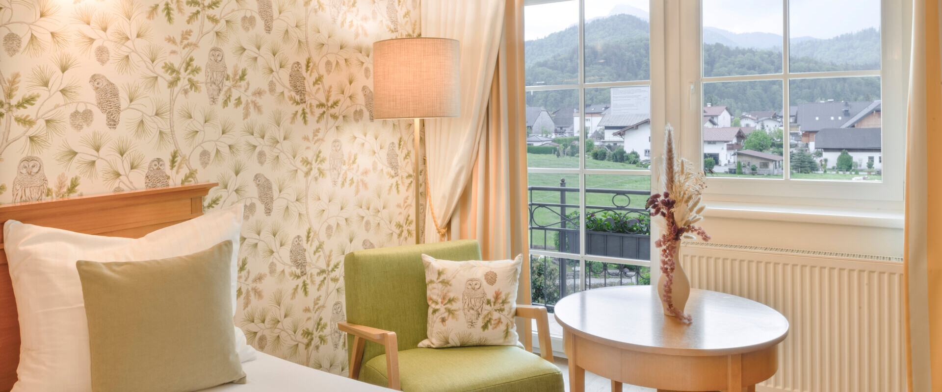 Cozy hotel room with floral wallpaper and a view of scenic mountains through a window.