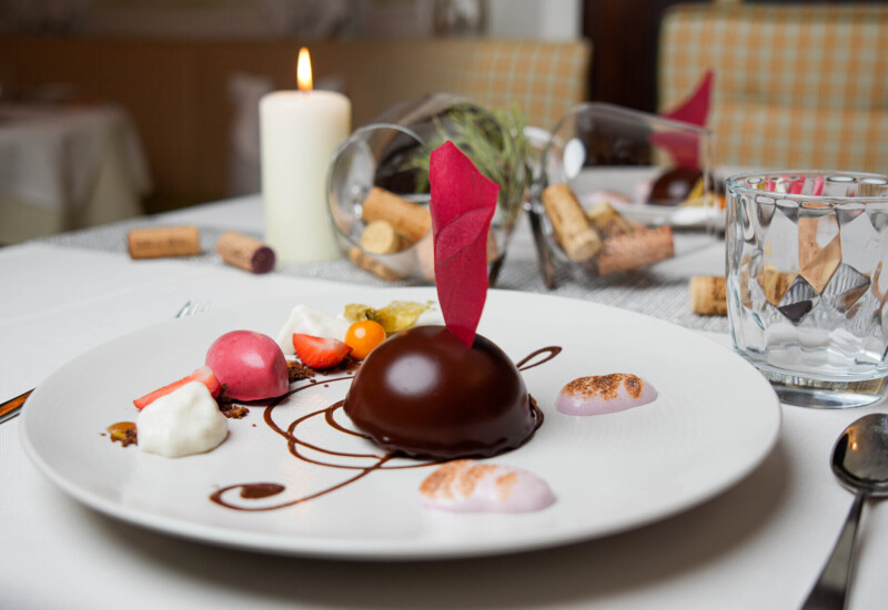 Gourmet dessert elegantly plated with colorful garnishes, set on a white tablecloth beside a candle and glass, creating a refined dining ambiance.