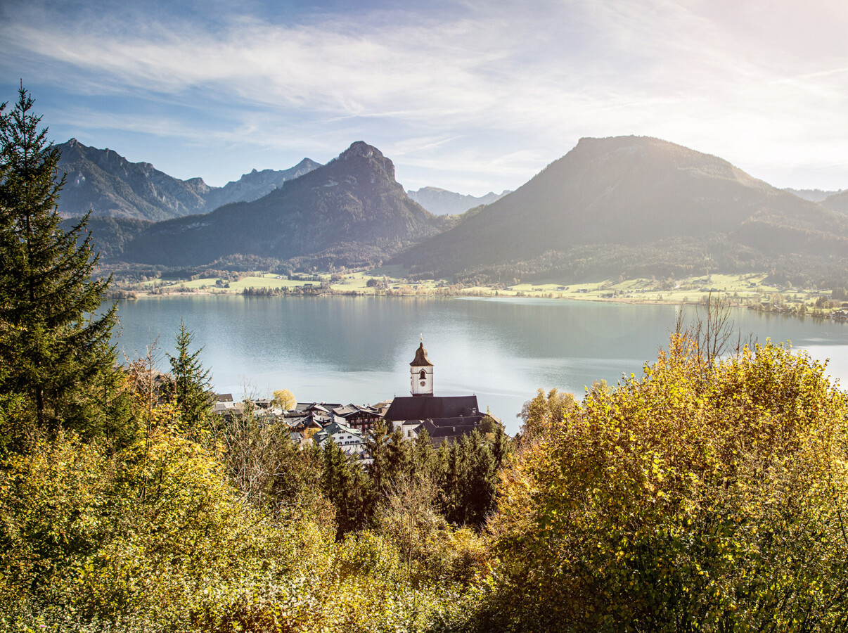 A scenic view of a serene lake surrounded by lush forests and mountains under a clear sky, with a small village in the foreground.