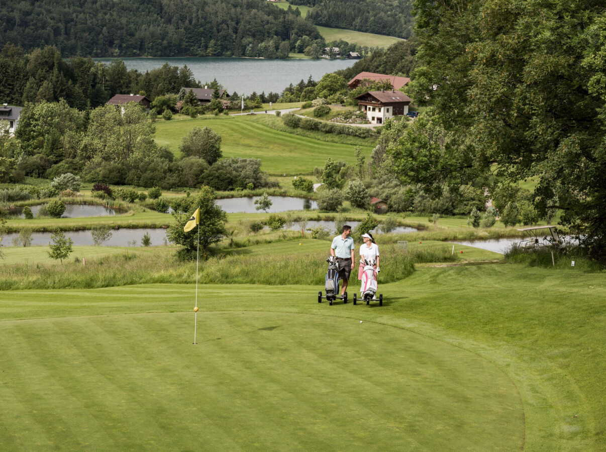 Two people walking on a golf course near Waldhof Fuschlsee Resort, with a flag in the foreground and a lake in the background.