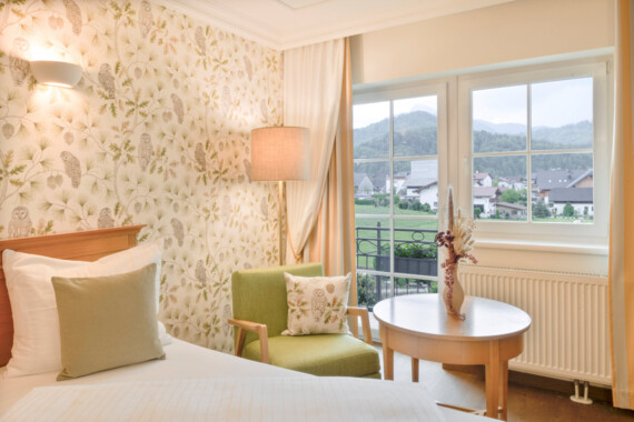Hotel room with floral wallpaper, a green chair, and a view of the countryside at Waldhof Fuschlsee Resort.