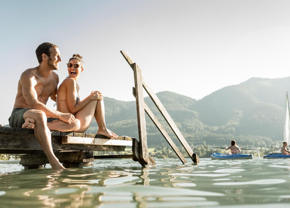 Couple enjoying a scenic view by a lake, sitting on a wooden dock, surrounded by lush mountains and people kayaking.