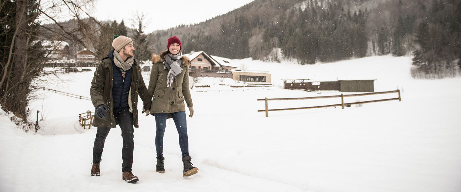 A couple walking in a snowy landscape with mountains and trees in the background.