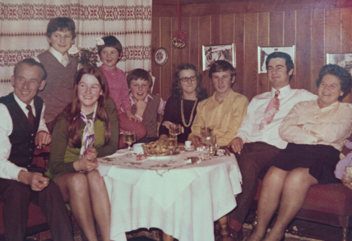 A family gathered around a table in a cozy, wood-paneled room, with drinks and a dessert on the table.