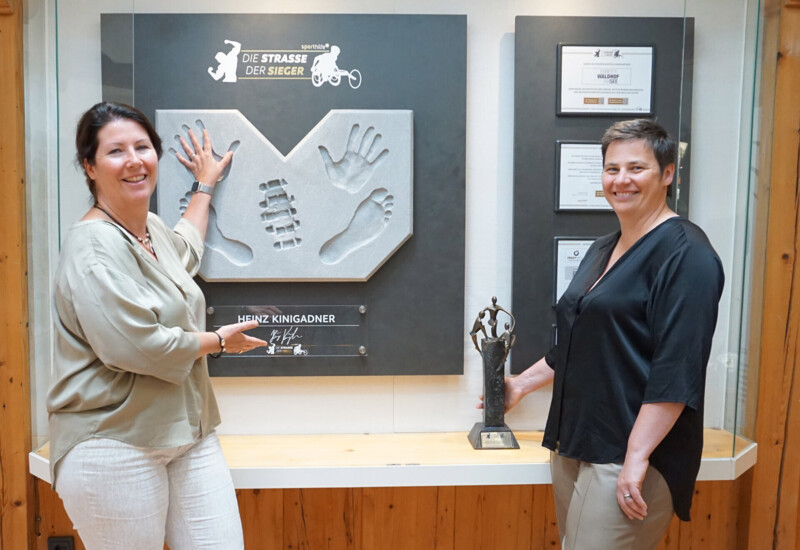 Zwei Frauen stehen lächelnd vor einer Wand mit Hand- und Fußabdrücken und einer Trophäe.